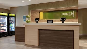 A hotel reception area with a wooden reception desk, a 'HOME 2' sign, and a 'Hilton Honors' sign. There are two beverage dispensers labeled 'really cold' and 'cold'.