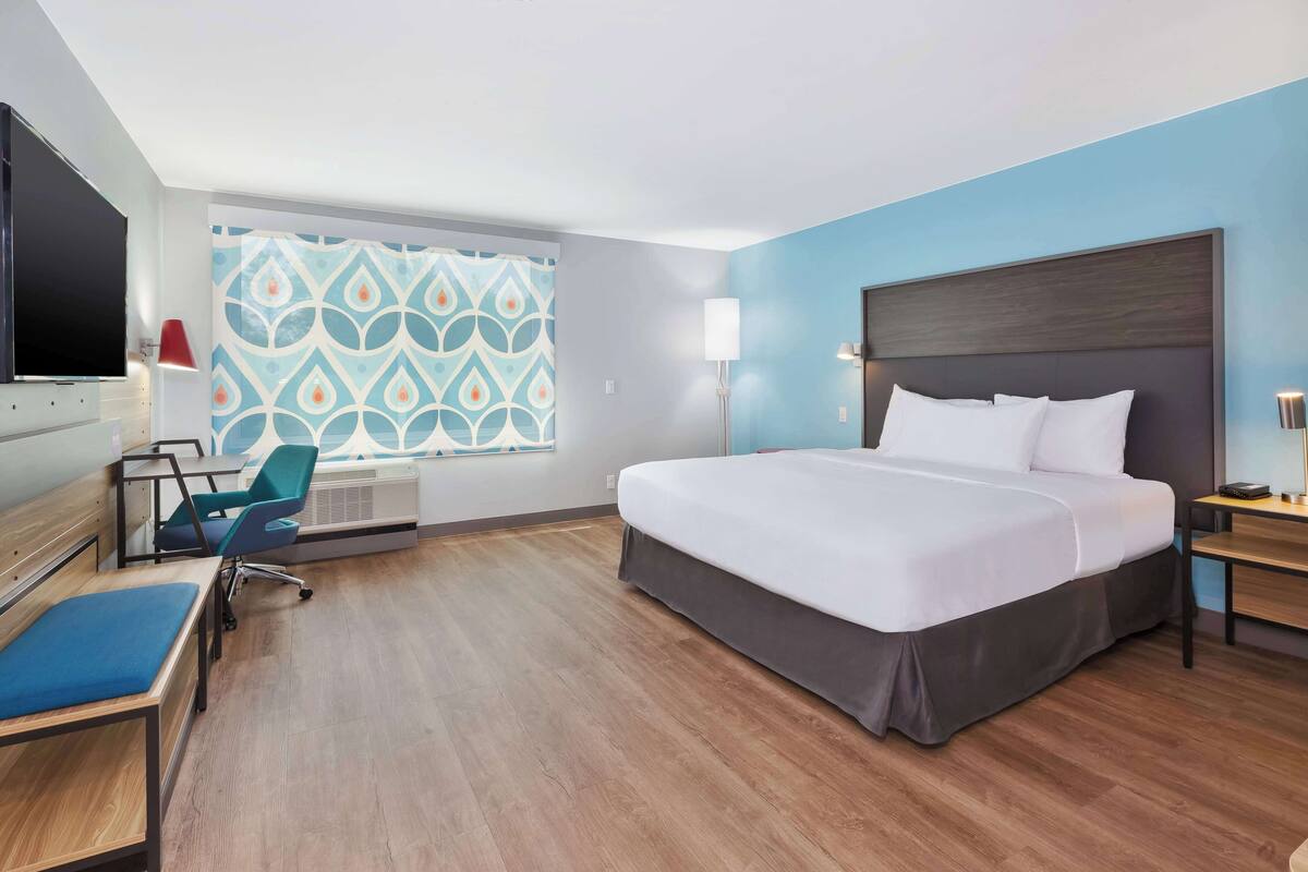 A hotel room with a large bed, a desk, a chair, a TV, and a window with patterned curtains.