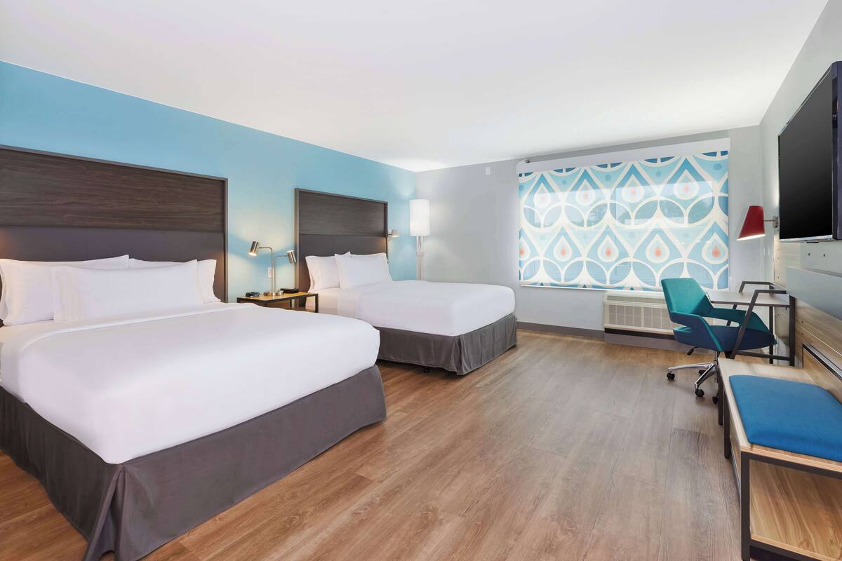 A hotel room with two beds, a desk, a chair, a TV, and a patterned wall.