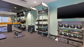 Et fitnessrum med træningsudstyr, et fjernsyn og en vægmonteret hylde med håndvægte og kettlebells.