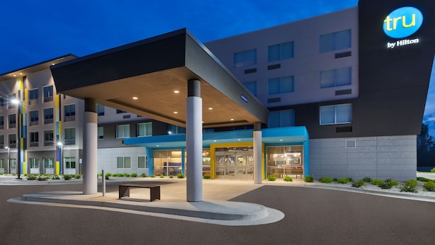Una entrada de hotel moderna con un área de espera cubierta, un llamativo letrero de Tru by Hilton y una fachada bien iluminada.