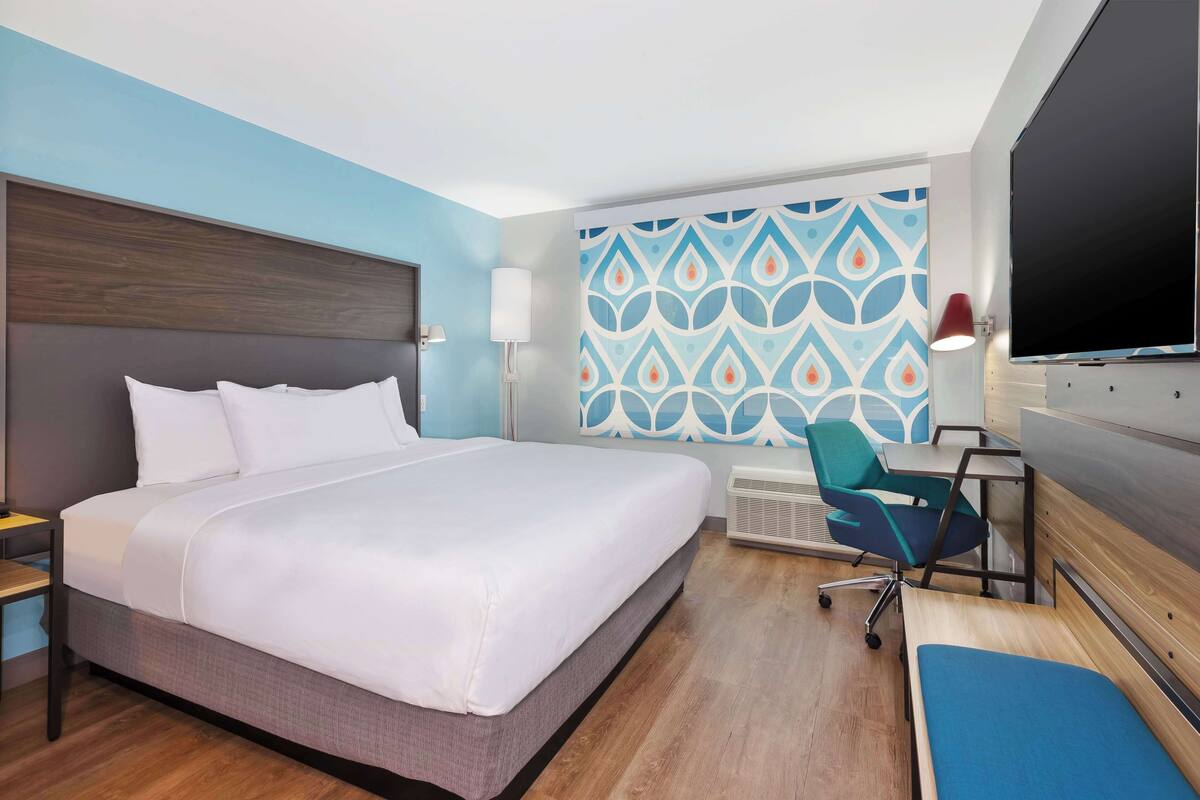 A hotel room with a large bed, a desk with a chair, a TV, and a patterned wall.