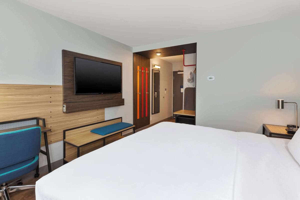A hotel room with a large bed, a desk with a chair, a TV mounted on the wall, and a shower area visible through an open door.