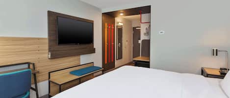 Quarto de hotel com uma cama grande, uma escrivaninha com cadeira, TV fixada na parede e zona de duche visível através de uma porta aberta.