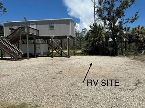 Une zone recouverte de gravier, avec un escalier en bois menant à une structure métallique, qui pourrait être une terrasse ou une plateforme, et une maison en arrière-plan.