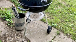 A black grill with a metal container and utensils, set on a concrete patio with grass and a wooden fence in the background. - Charm Near Omaha Downtown: 4-Bed, 2-Bath Gem (Omaha)