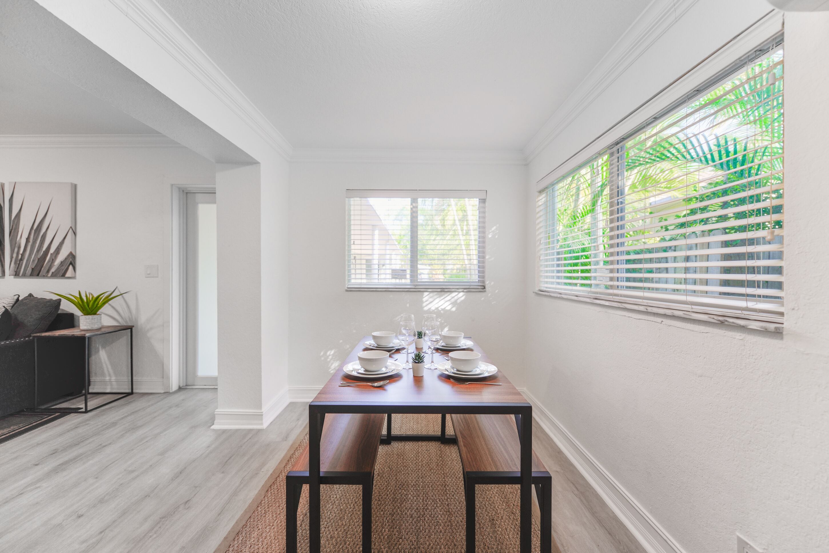 A dining area with a wooden table set for six, white walls, and a window with blinds.