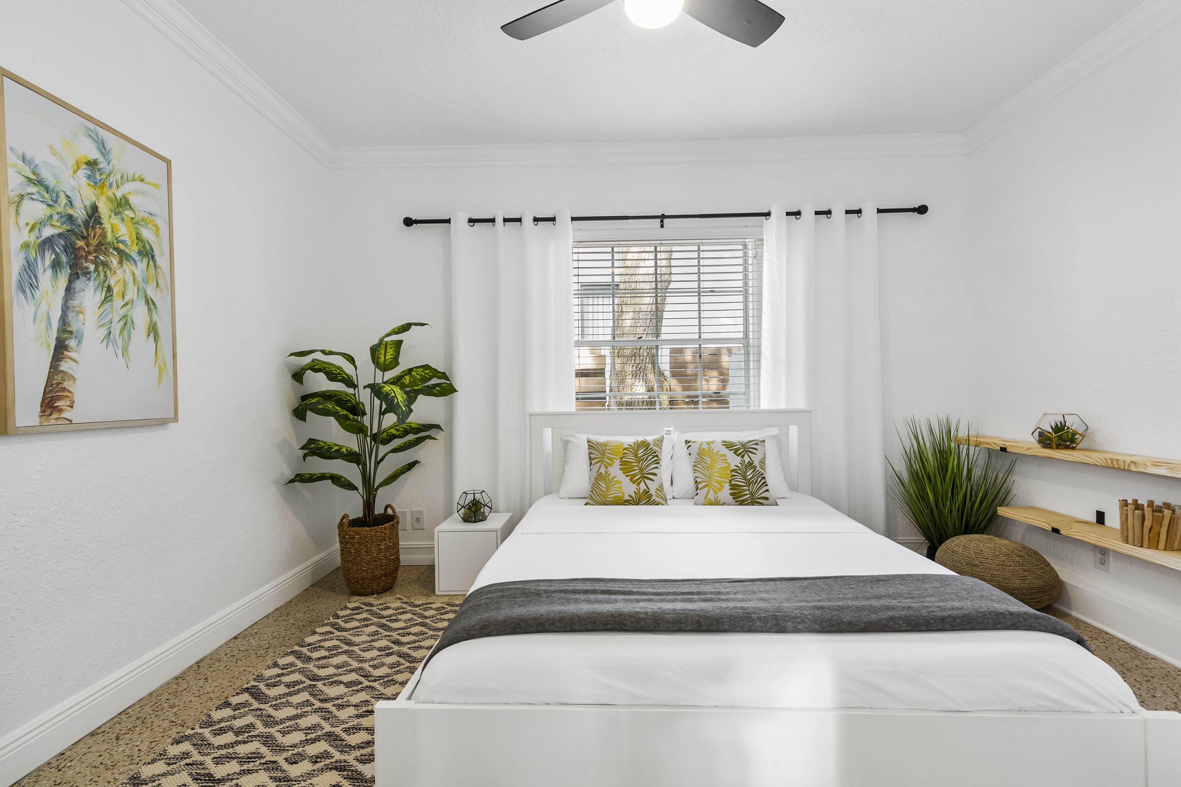 A bedroom with a bed, a window with blinds, a ceiling fan, a potted plant, and a wall-mounted shelf.