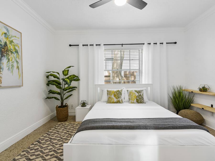 A bedroom with a bed, a window with blinds, a ceiling fan, a potted plant, and a wall-mounted shelf.