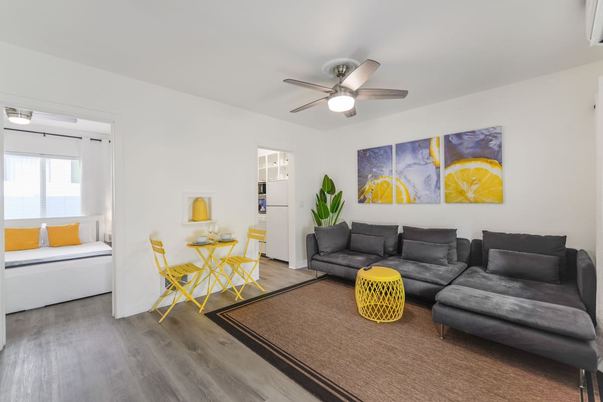 Ein modernes Wohnzimmer mit grauem Sofa, gelben Sesseln, einem kleinen Tisch und Kunstwerken an der Wand.