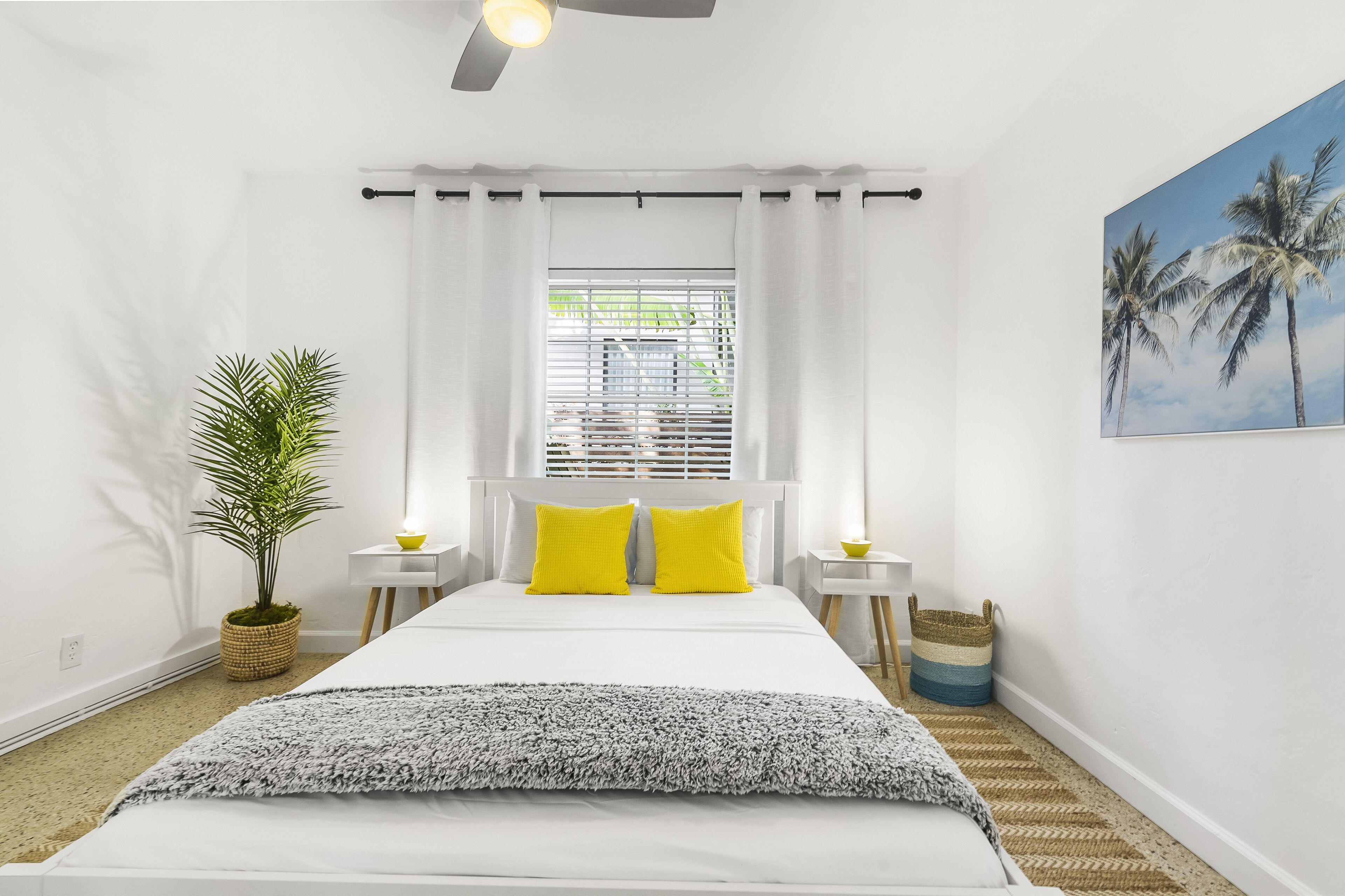 Un dormitorio con cama, cortinas blancas, ventilador de techo, ventana con persianas, planta en maceta, una mesita de noche con lámpara y un cuadro en la pared.