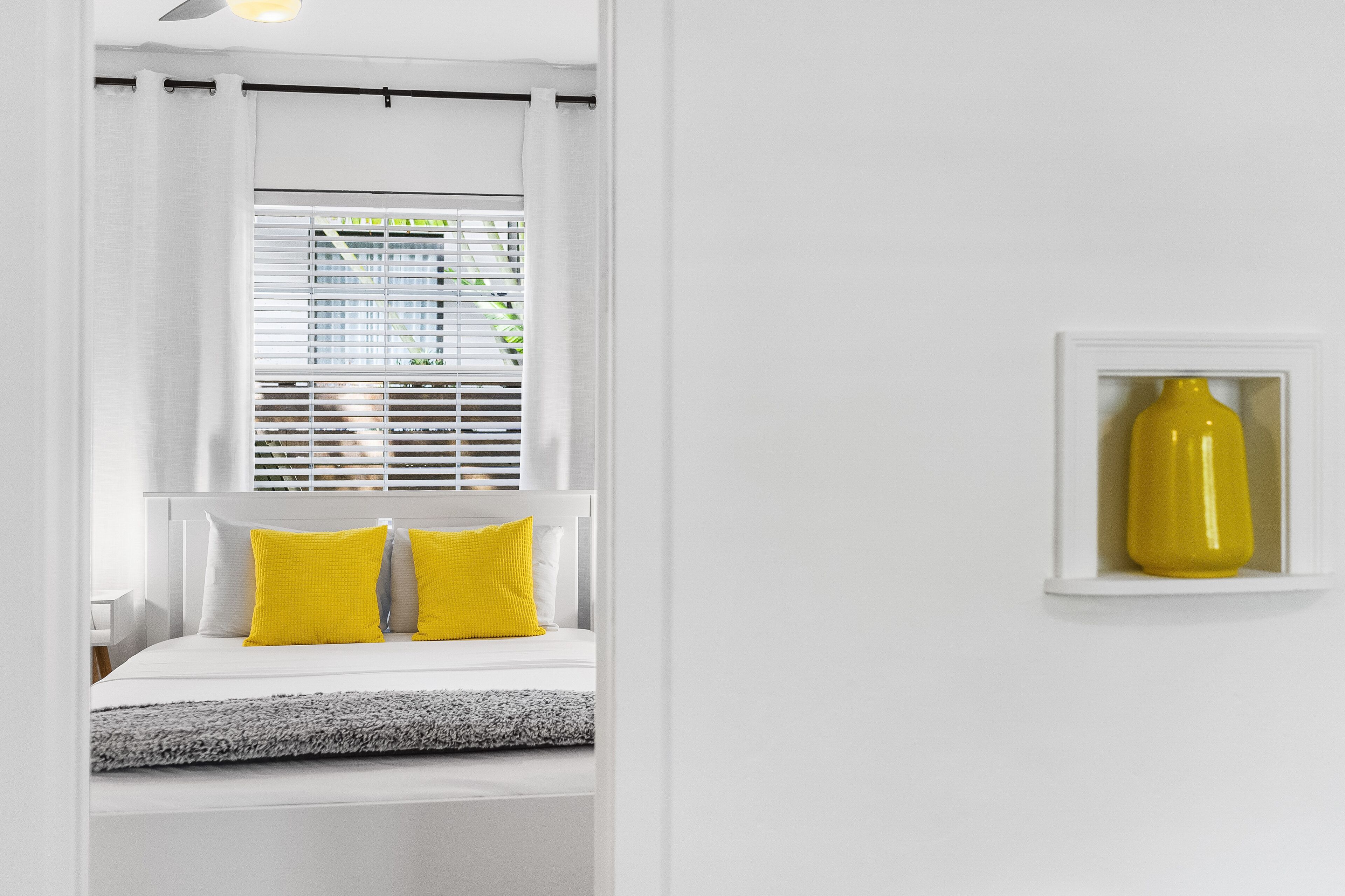 A bedroom with a bed, white curtains, a window with blinds, and a yellow vase on a shelf.