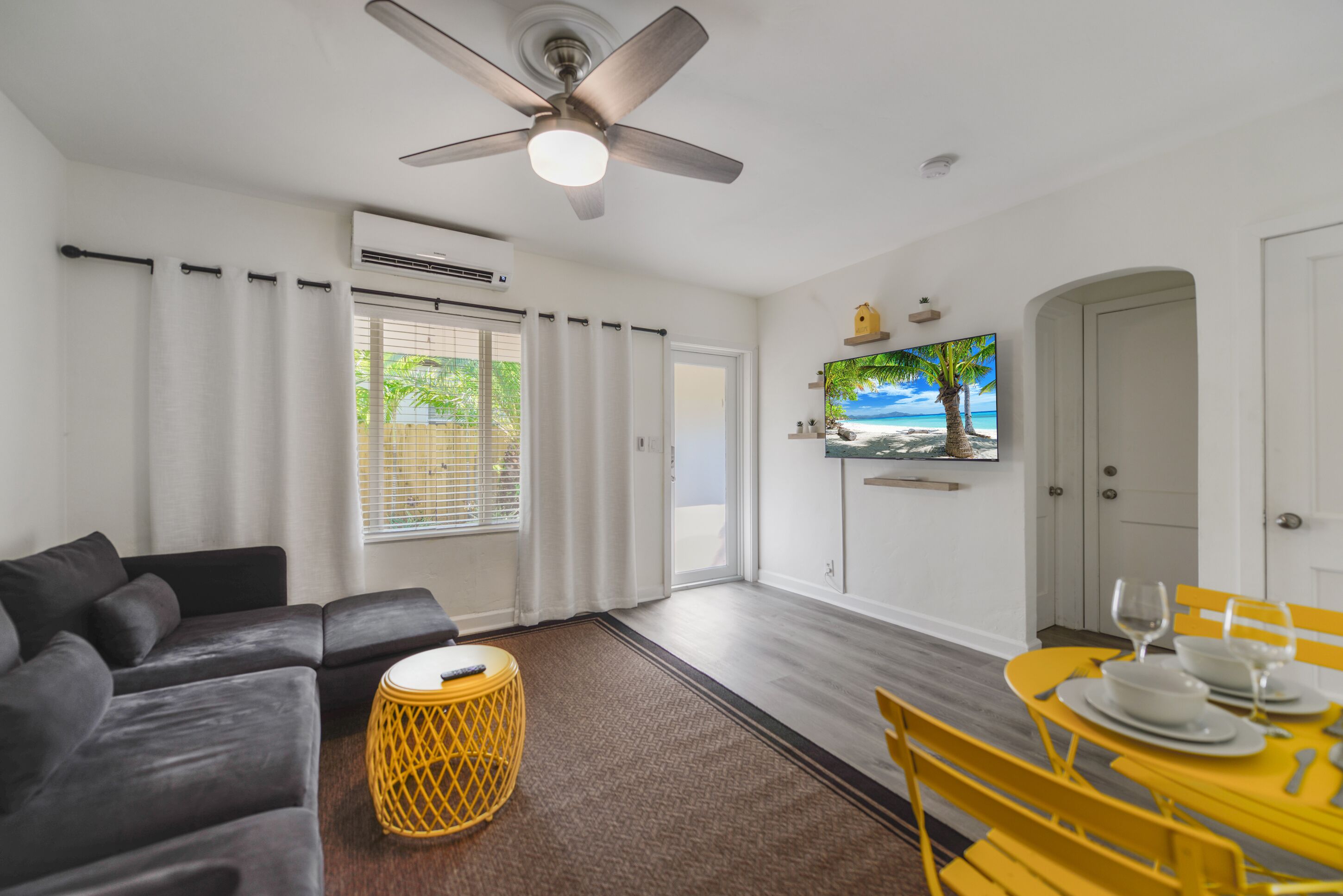 A modern living room with a sofa, a dining table set for two, a ceiling fan, and a TV mounted on the wall.