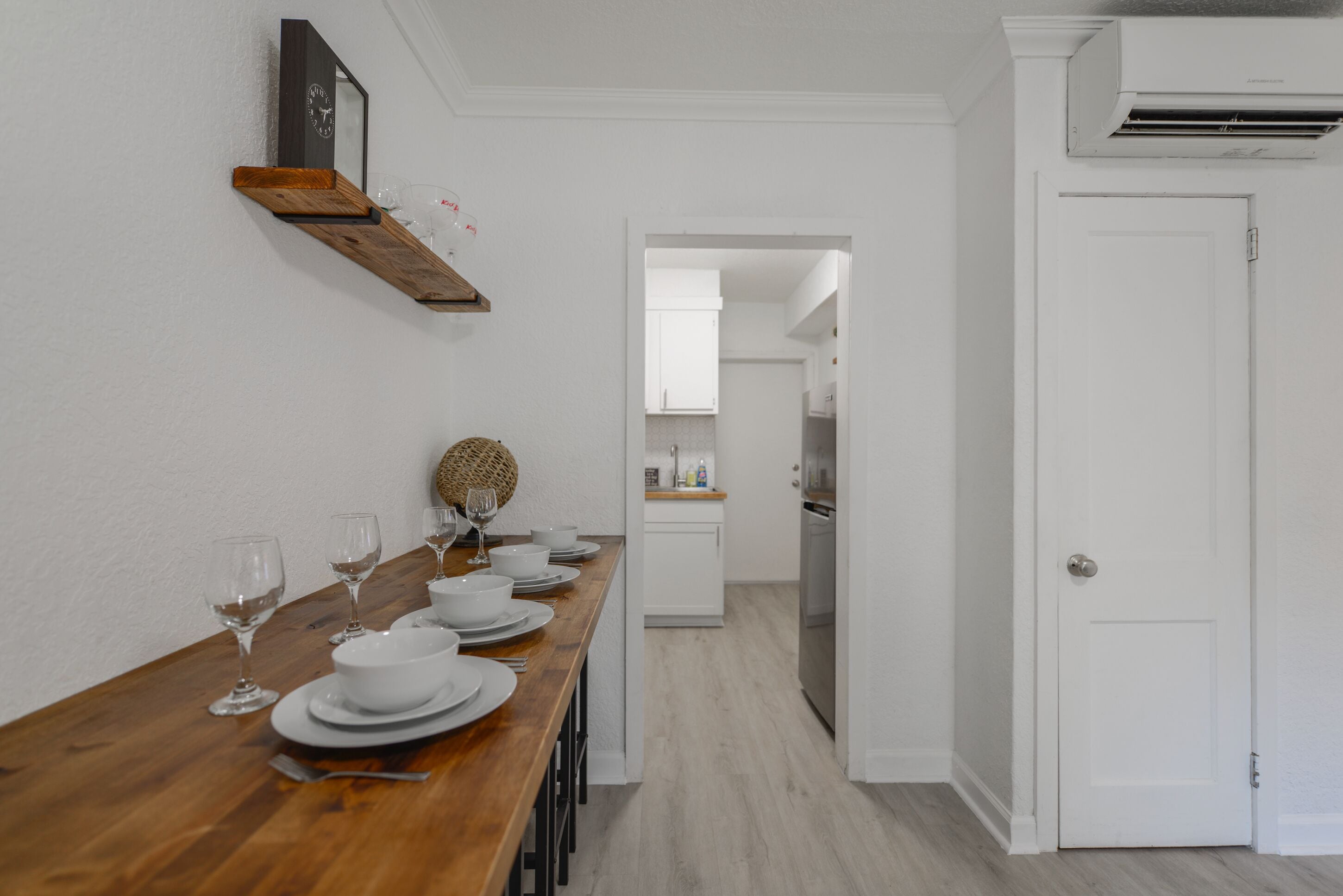 A dining area with a wooden table set for a meal, a kitchen in the background, and a white door.