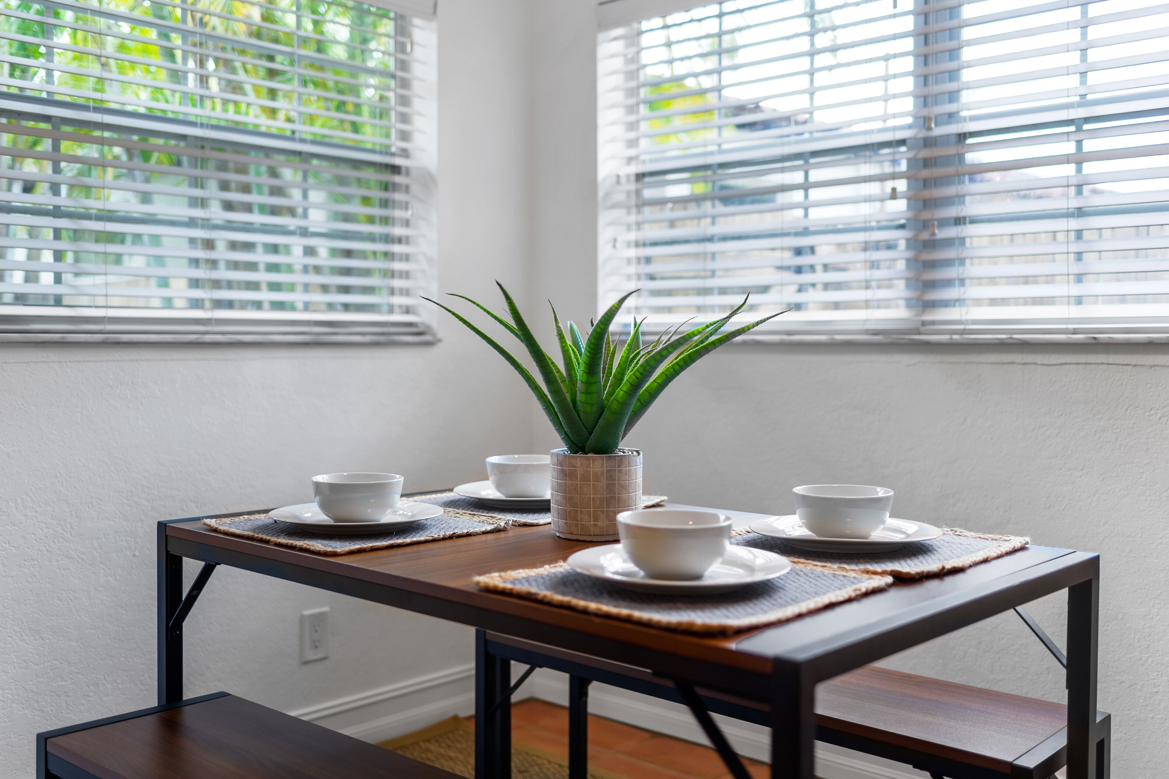 A dining table with white bowls and a potted plant, set against a white wall with blinds.