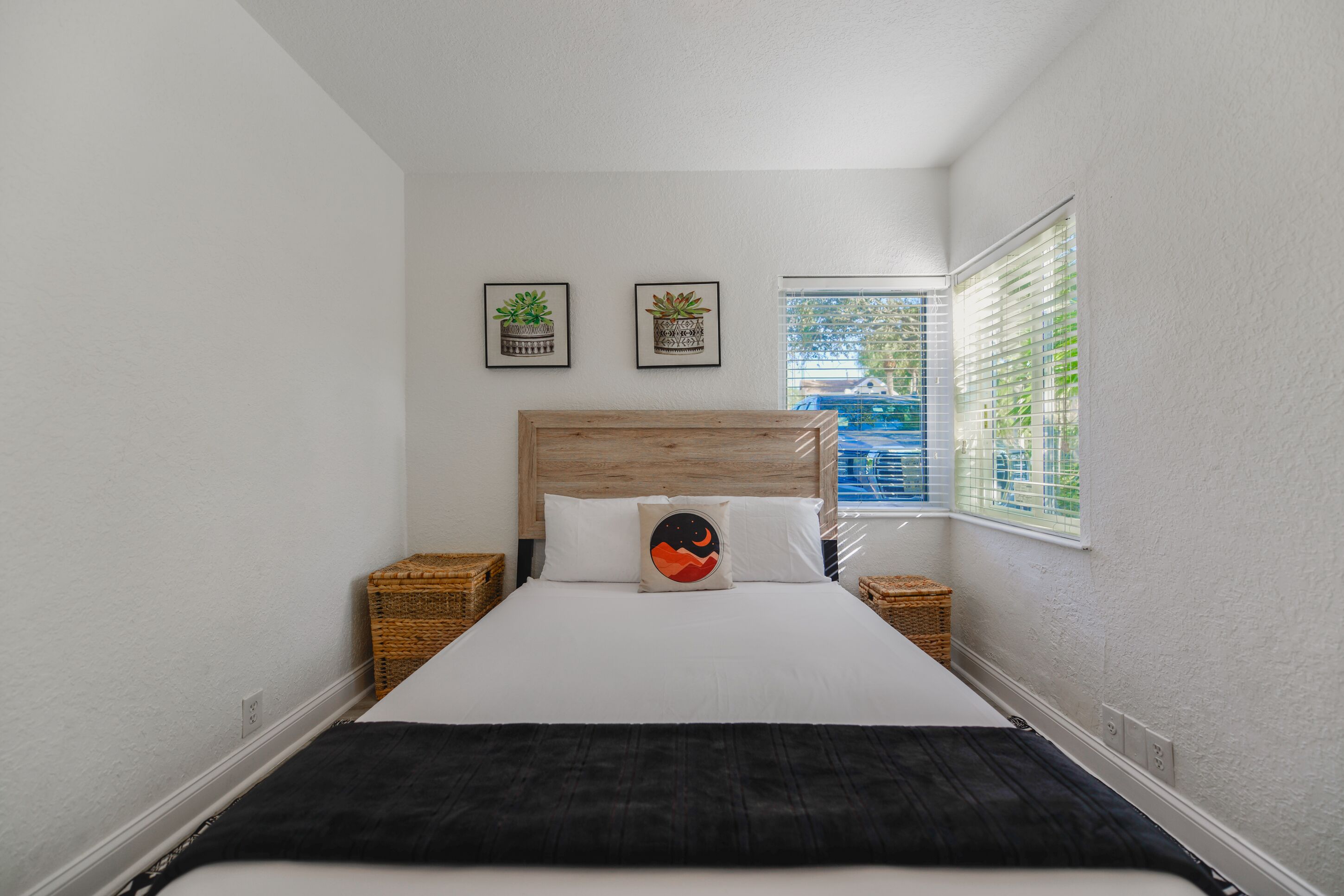 A neatly made bed with a wooden headboard, two framed pictures on the wall, and a window with blinds.