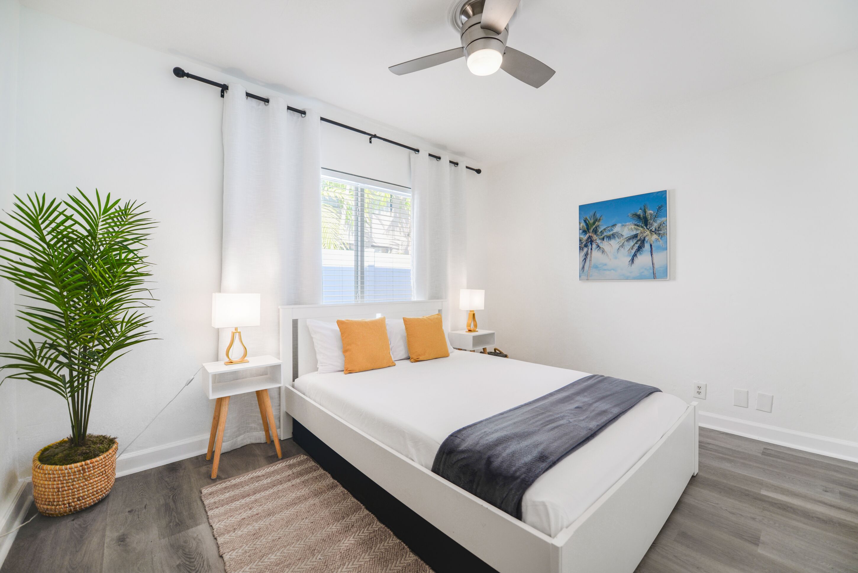 Un dormitorio moderno con una cama grande, paredes blancas, un ventilador de techo, una ventana con cortinas, una planta en maceta y un cuadro de palmeras.