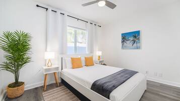 Un dormitorio moderno con una cama grande, paredes blancas, un ventilador de techo, una ventana con cortinas, una planta en maceta y un cuadro de palmeras.