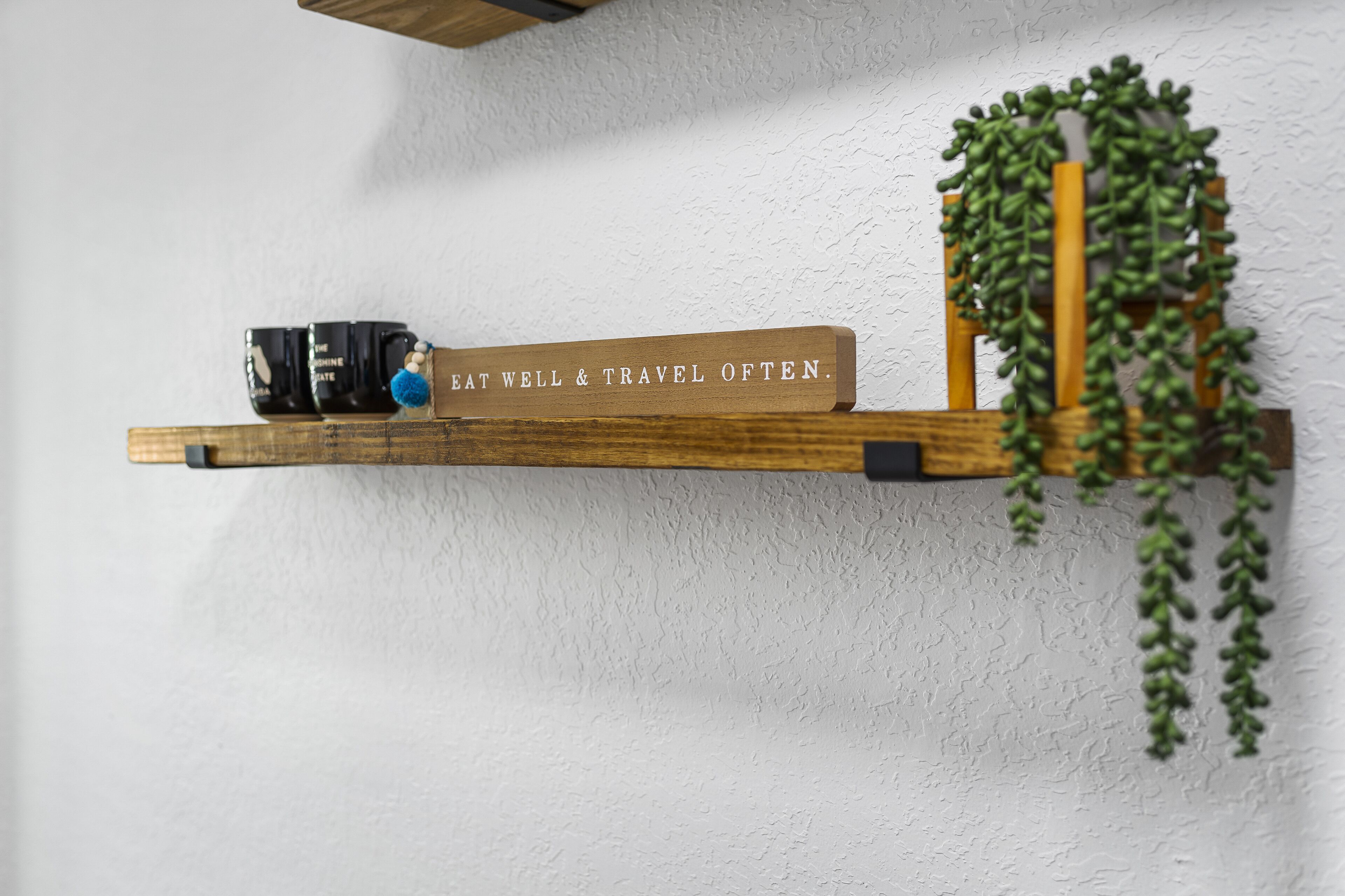 A wooden shelf with a motivational sign, a mug, and a hanging plant against a textured wall.
