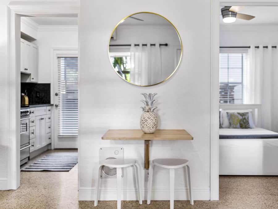 A modern, white-themed room with a round mirror, a wooden table, two white chairs, and a bed with a white headboard.