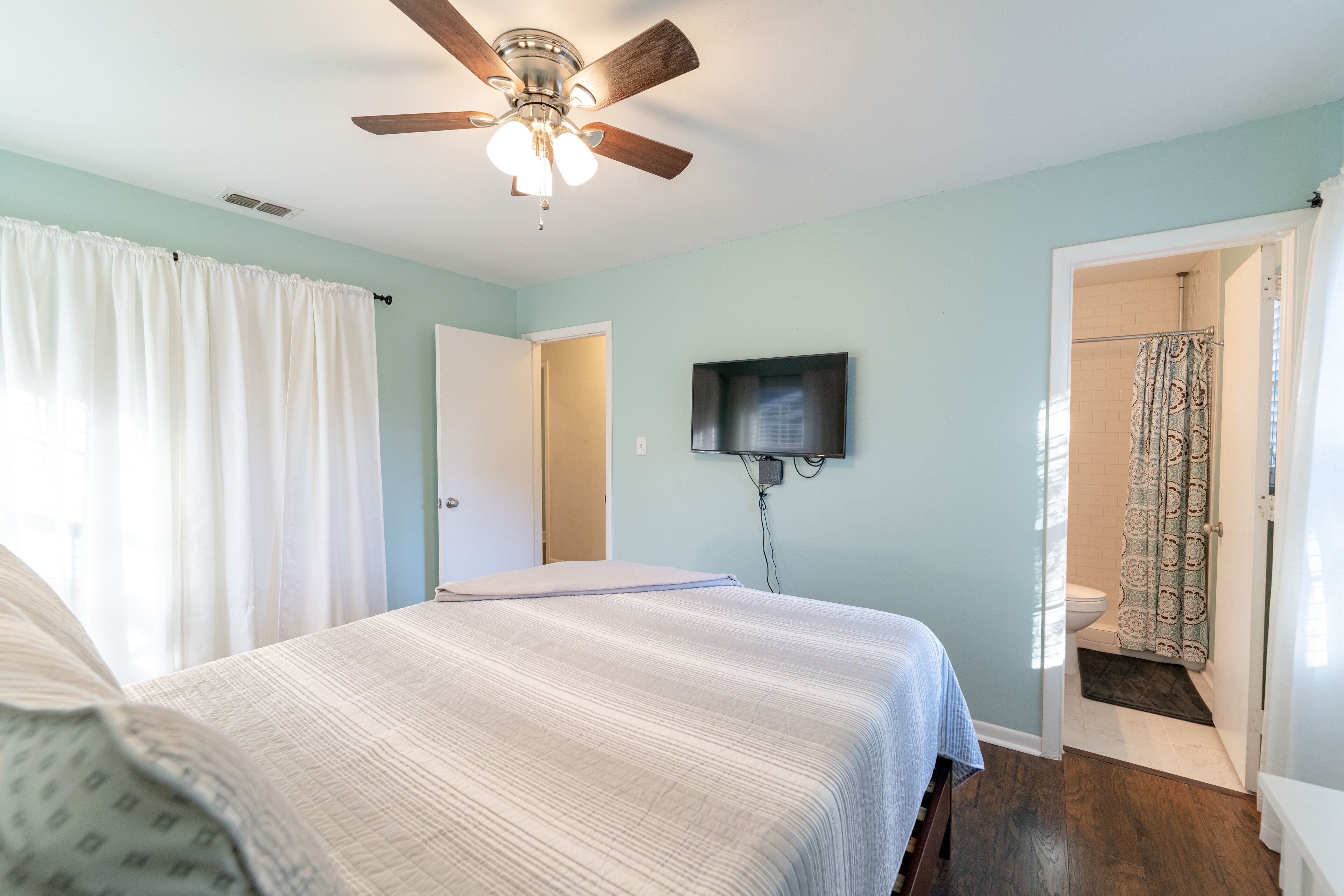 Une chambre à coucher avec un lit, un ventilateur de plafond, un téléviseur fixé au mur et une salle de bain visible à travers une porte ouverte.