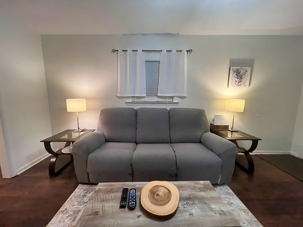 Ein Wohnzimmer mit grauem Sofa, zwei Beistelltischen mit Lampen, einem Couchtisch mit Hut und Fernbedienungen sowie einem Fenster mit Vorhängen.