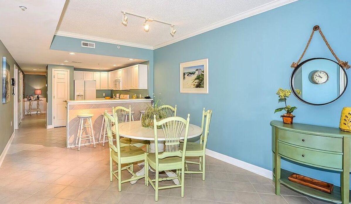 A dining area with a round table and chairs, a kitchen in the background, and a green console table with a plant.