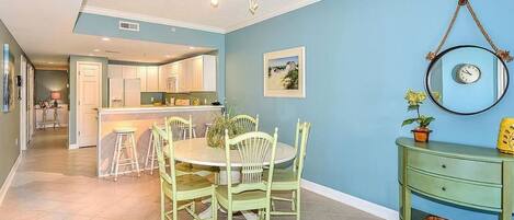 A dining area with a round table and chairs, a kitchen in the background, and a green console table with a plant.