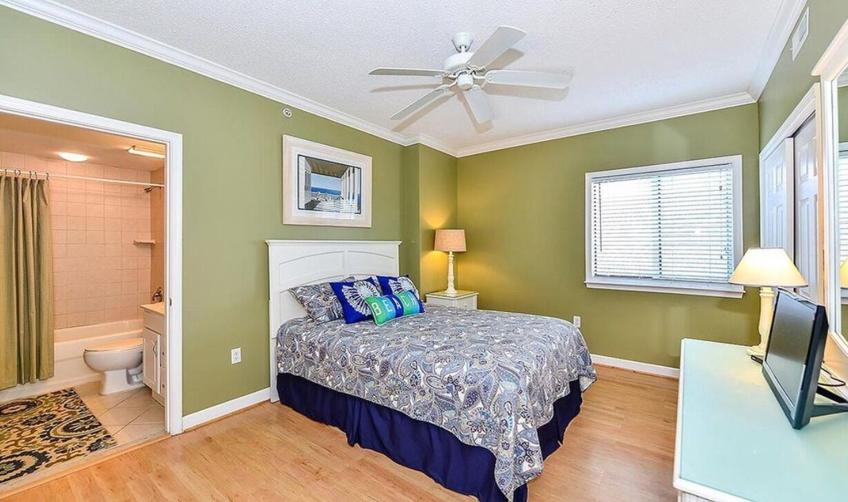 A bedroom with a bed, a ceiling fan, a window with blinds, a bedside table with a lamp, and a bathroom with a shower and toilet.