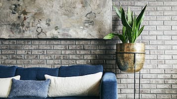A blue sofa with patterned cushions against a brick wall, a textured painting above, and a hanging plant in a textured pot.