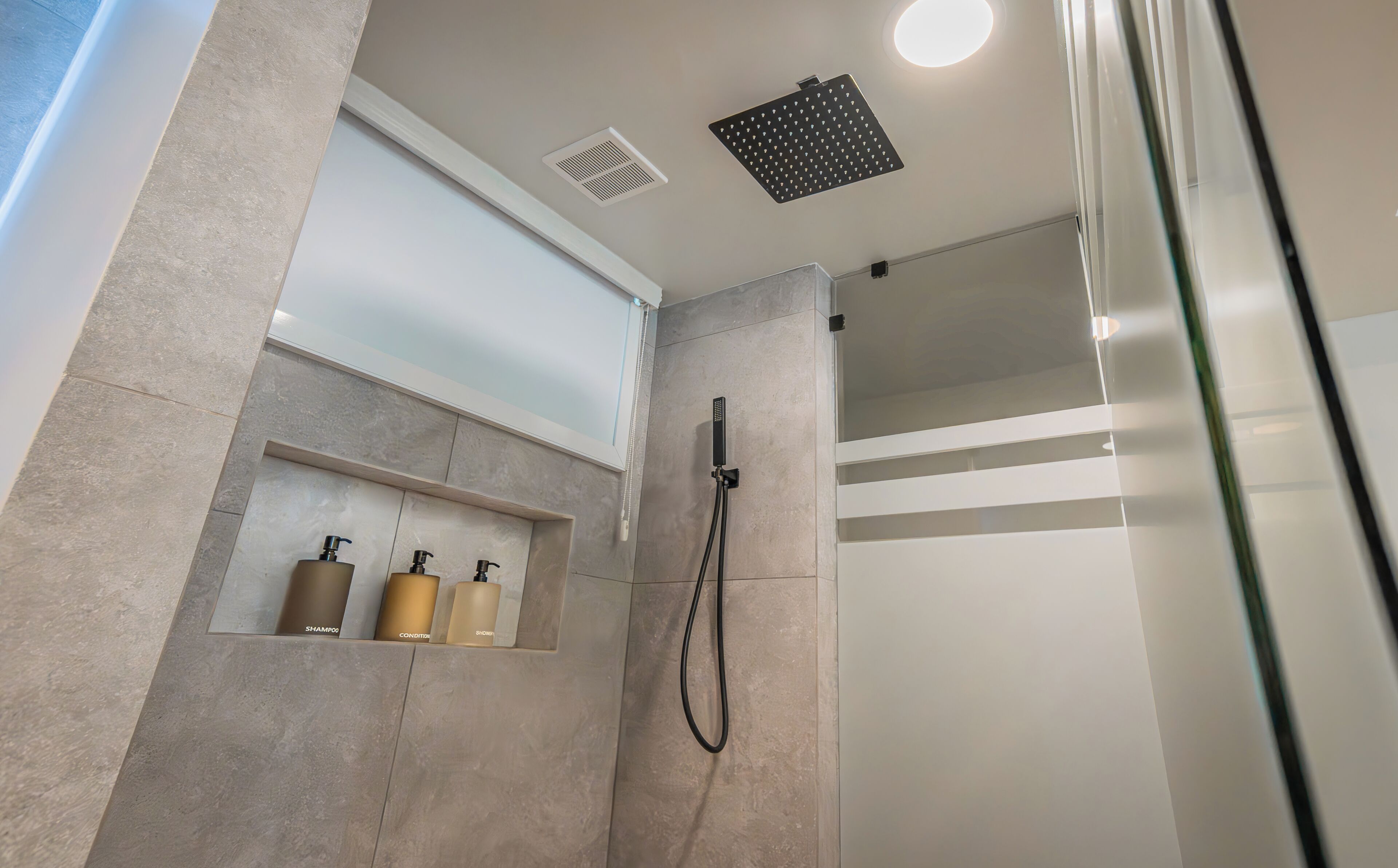 A modern shower area with a handheld showerhead, a built-in shelf with toiletries, and a ceiling vent.
