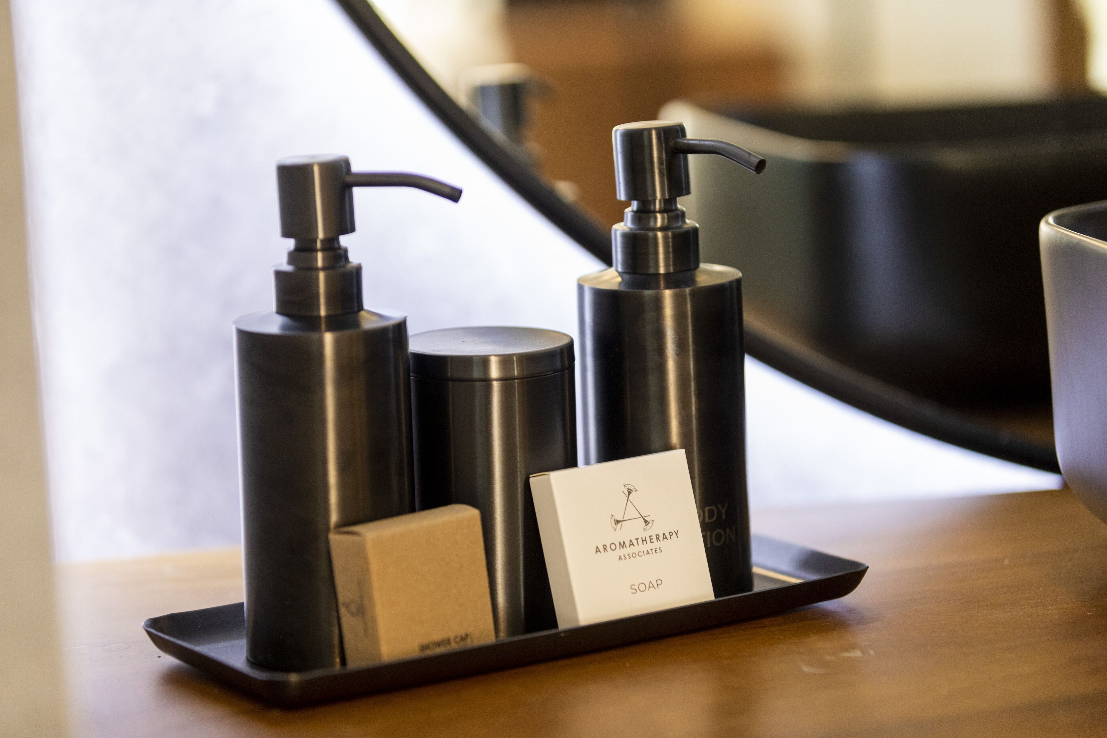 A tray with two black dispensers and a soap bar labeled "Aromatherapy Associates Soap" on a wooden surface.