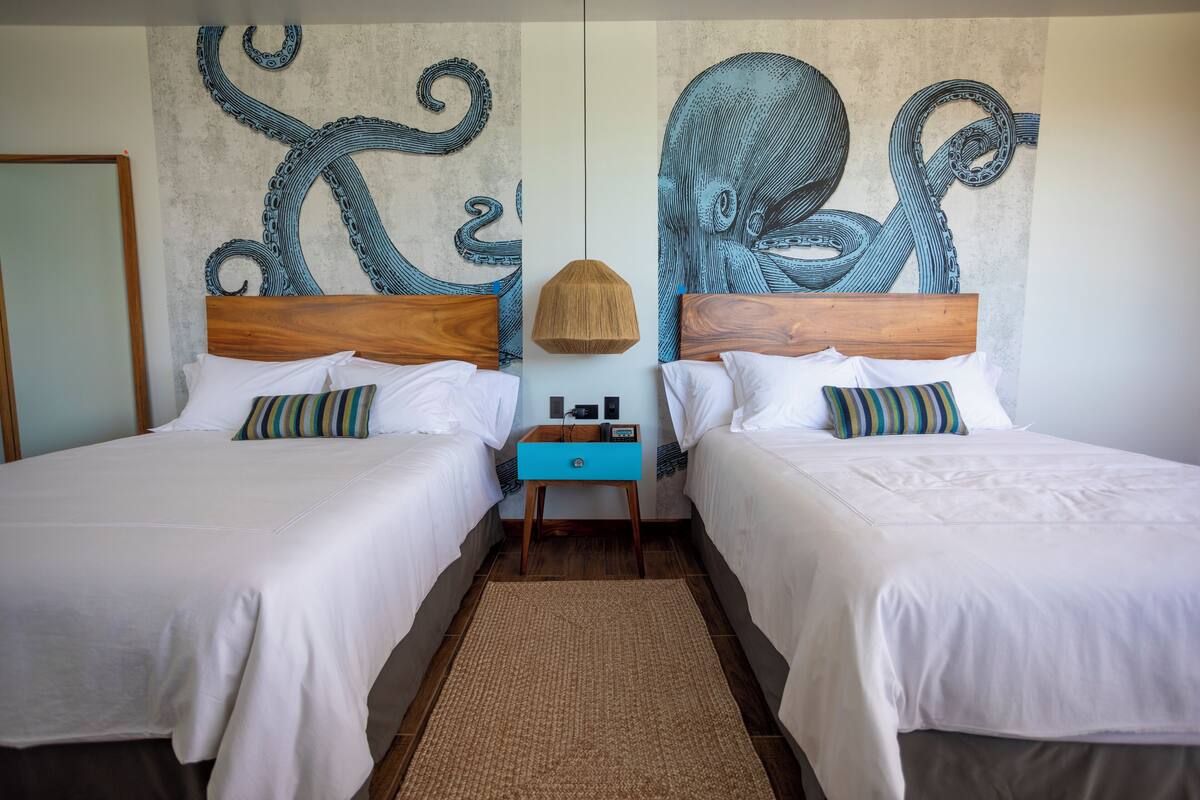 Habitación de hotel con dos camas, un cabecero de madera, una mesita de noche azul y un mural grande con forma de pulpo en la pared.