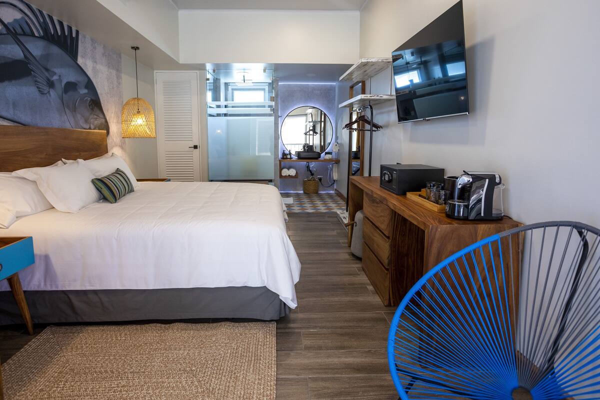 Habitación de hotel con cama, televisión, escritorio y cafetera, y baño visible al fondo.