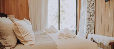 A bedroom with a large bed, white linens, a stone wall, and a window with a view.