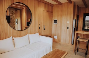 Een kamer met een houten wand, een ronde spiegel, een witte bank, een klein houten tafeltje en een koelkast.