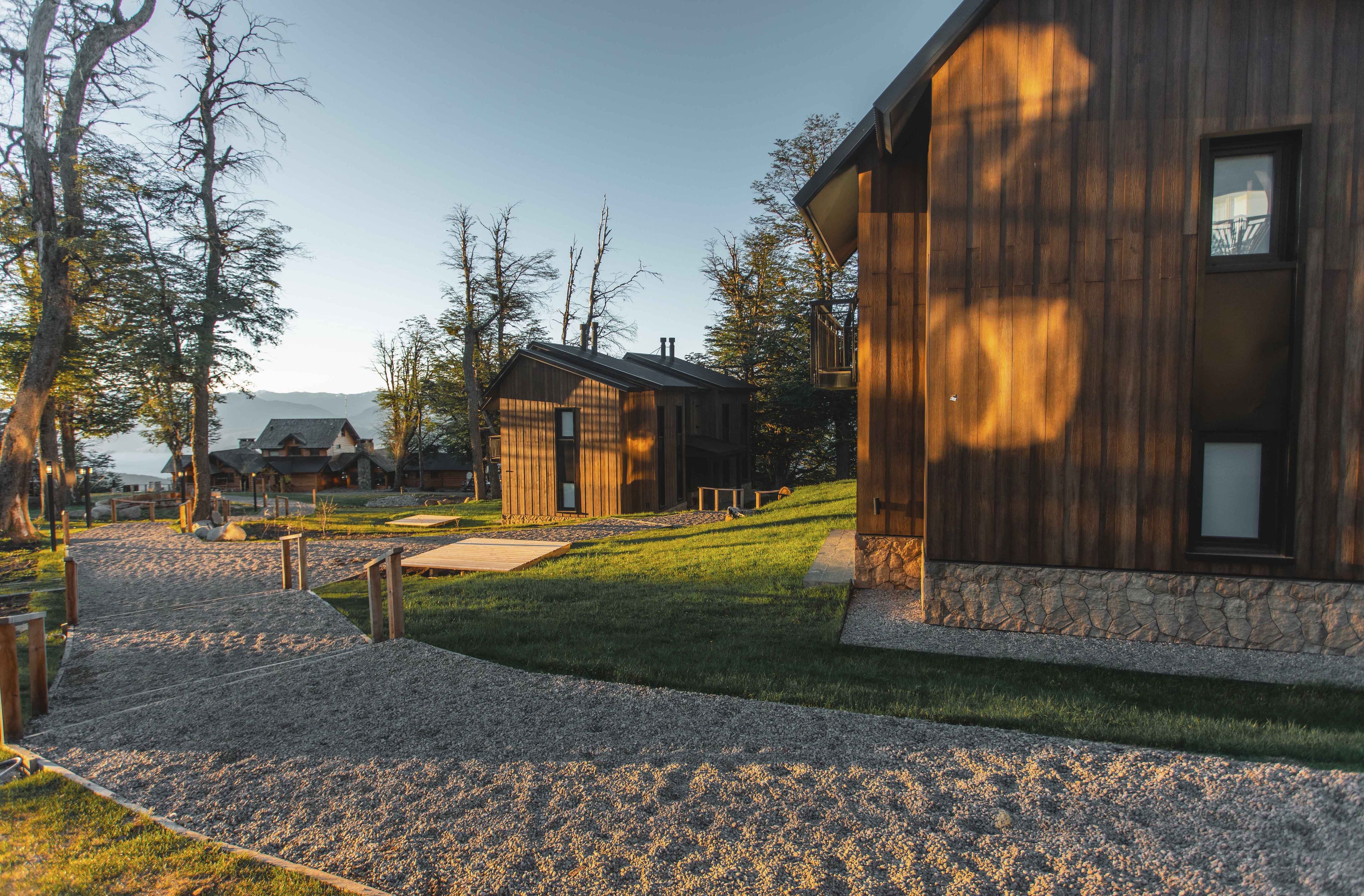 Une cabane moderne en bois, dotée de fondations en pierre, entourée d’un chemin de gravier et de zones herbeuses, et située dans un paysage boisé.