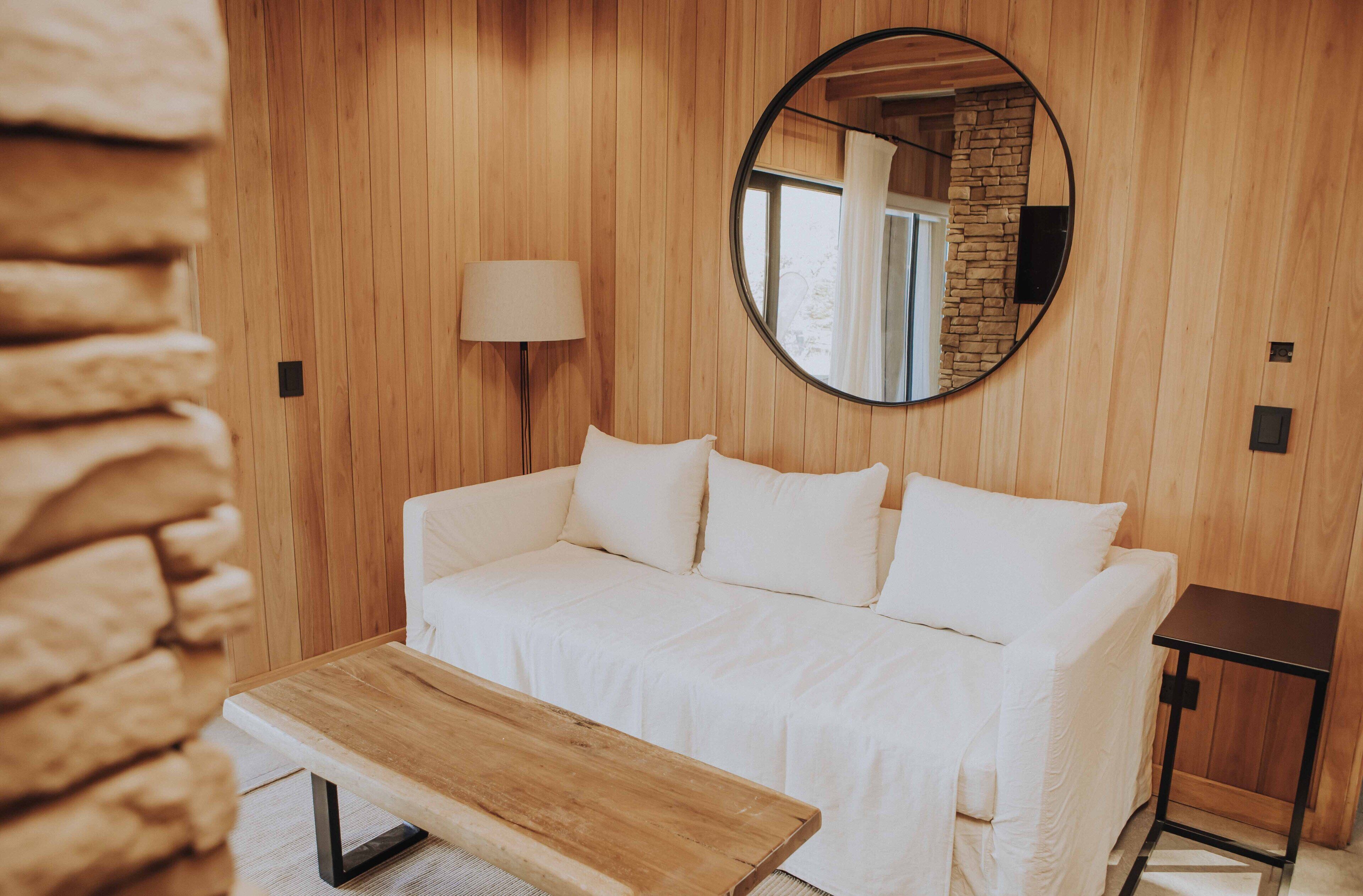 Una habitación con paneles de madera, un sofá blanco, un espejo redondo y una pequeña mesa.