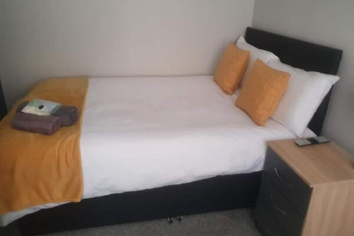 A bed with a white mattress, two orange pillows, and a brown blanket. There is a wooden nightstand with a drawer and a small item on top.