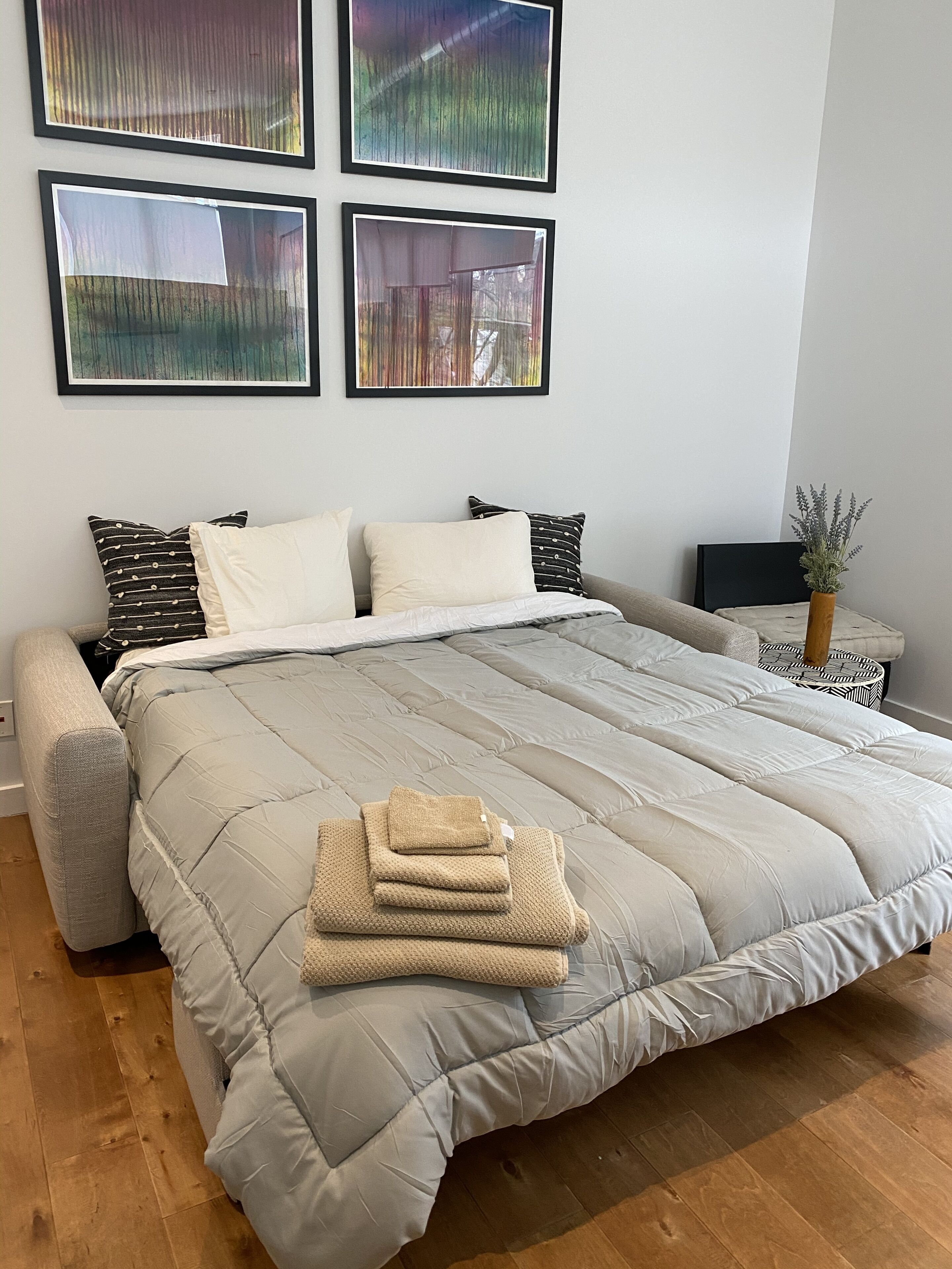A neatly made bed with a grey quilt, a bedside table with a plant, and abstract art on the wall.