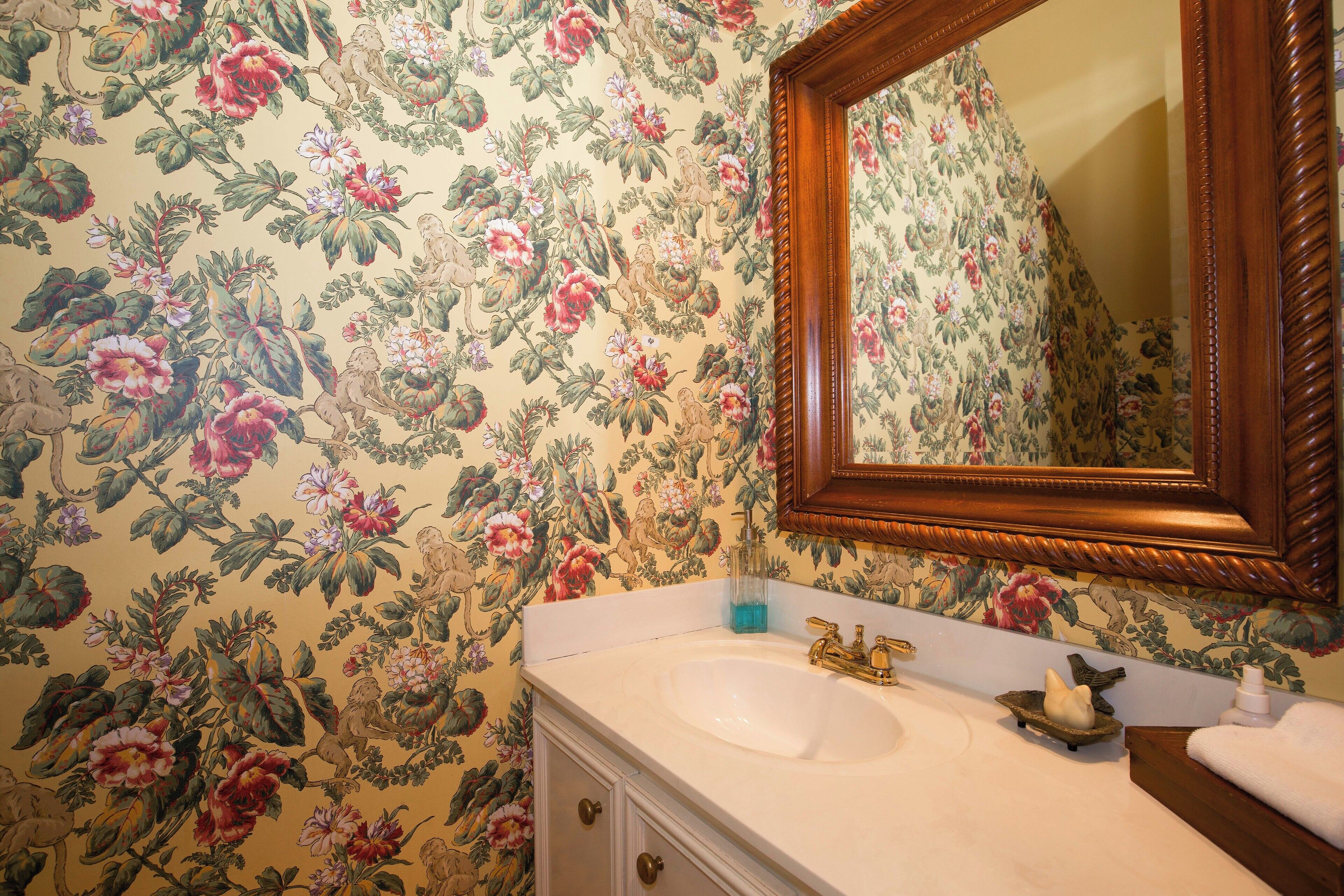Une salle de bain avec un papier peint à motifs floraux, un miroir encadré en bois, un lavabo blanc et un petit oiseau décoratif.