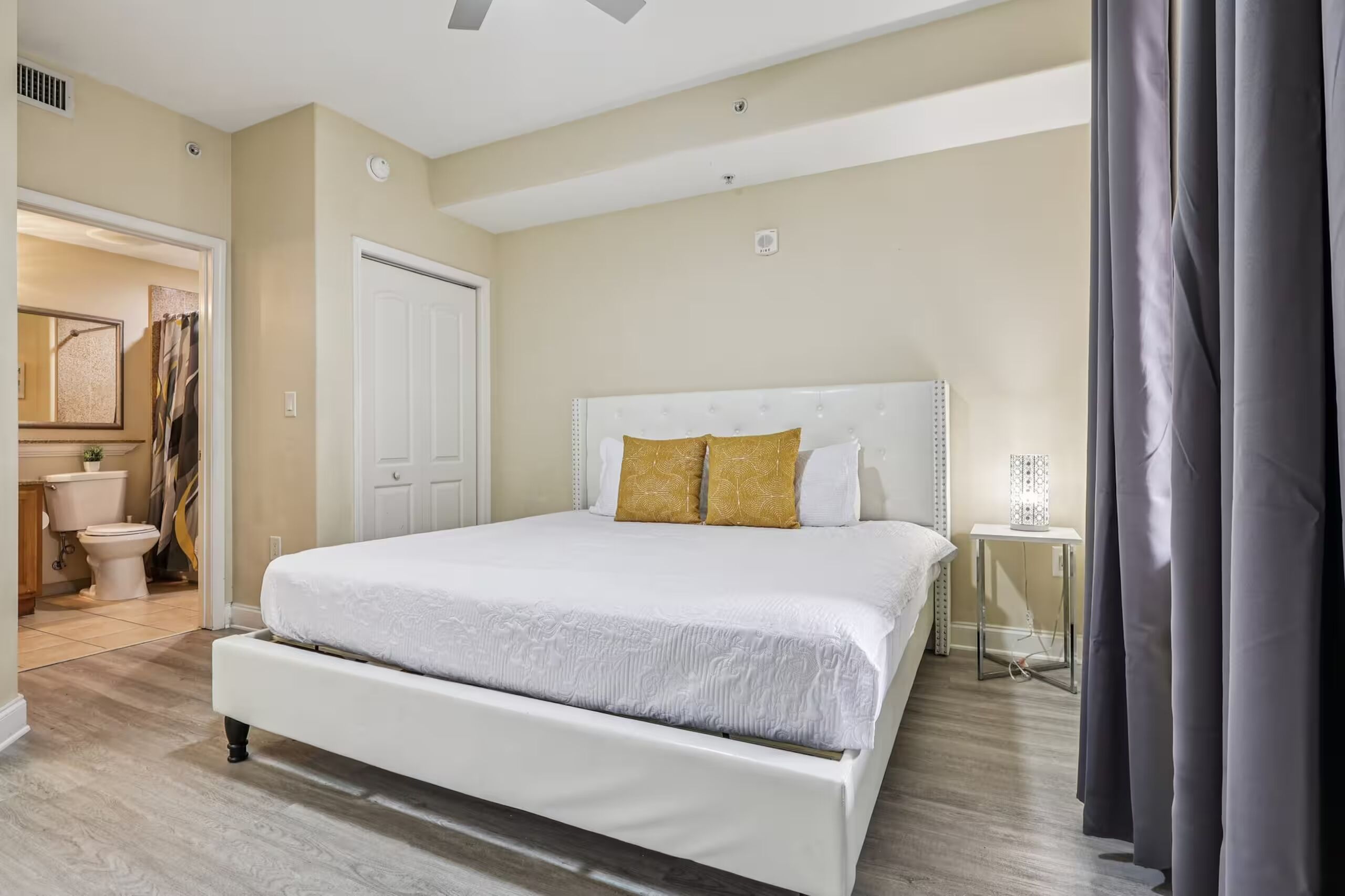 A bedroom with a large bed, a white door, a small table with a lamp, and a view of the bathroom.
