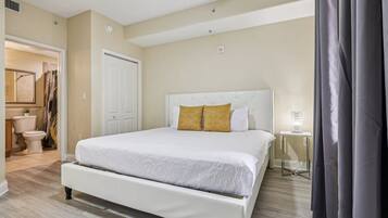 A bedroom with a large bed, a white door, a small table with a lamp, and a view of the bathroom.