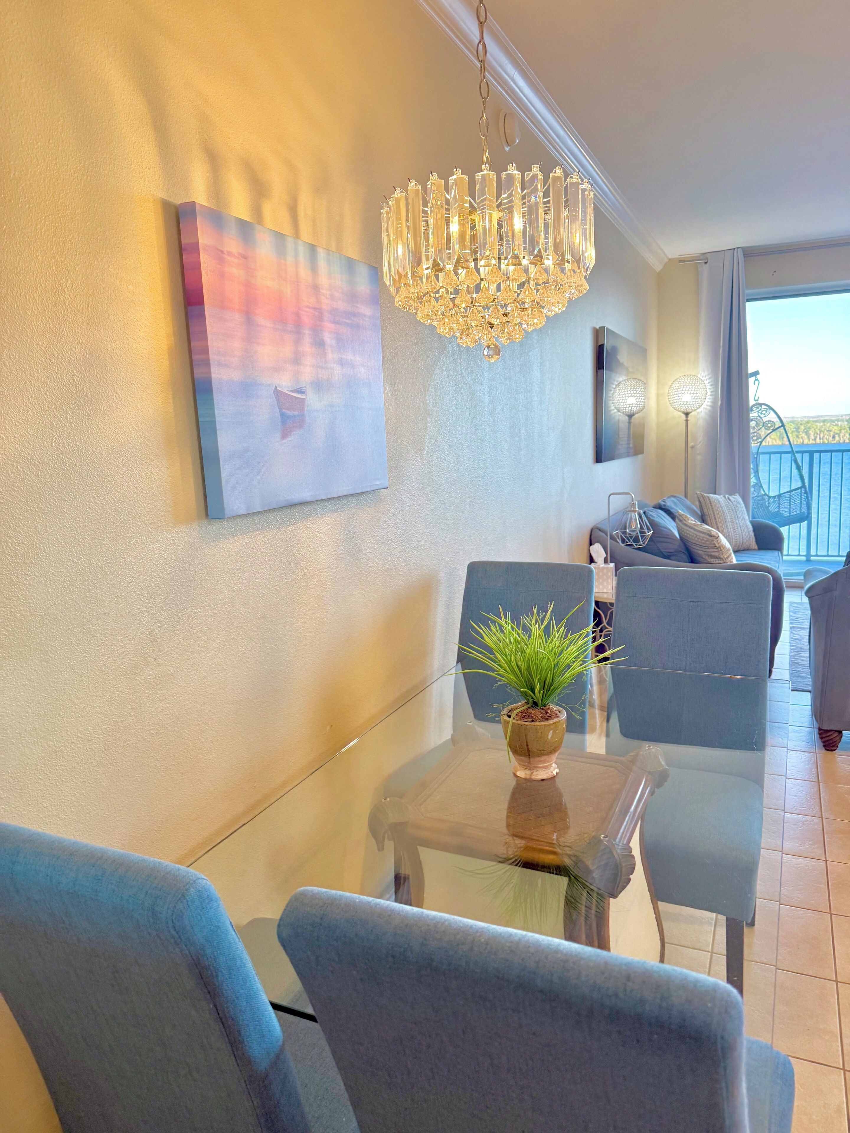 Un salon moderne avec une table en verre, des chaises bleues, un lustre, un tableau et un balcon avec vue.