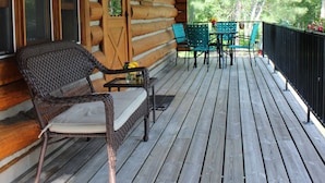 Een houten blokhut met een veranda, voorzien van rieten stoelen en een tafel, omringd door een zwarte hekwerk.