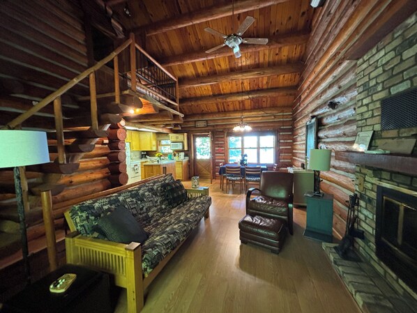A log cabin interior with a wooden staircase, a sofa, a leather chair, a fireplace, and a dining area.