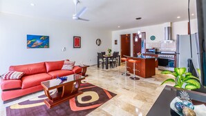 A modern living room with a red sofa, a glass coffee table, and a dining area with a kitchen in the background.