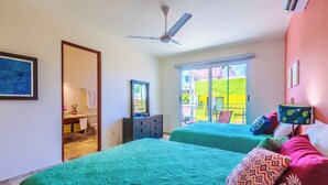 A bedroom with two beds, a ceiling fan, a desk with a chair, and a window with a view of a colorful building.