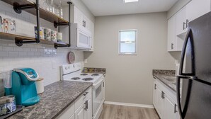 Una cocina compacta con armarios blancos, un refrigerador negro, una estufa y un microondas. Hay una ventana sobre el lavamanos y una repisa con cristalería y recipientes.