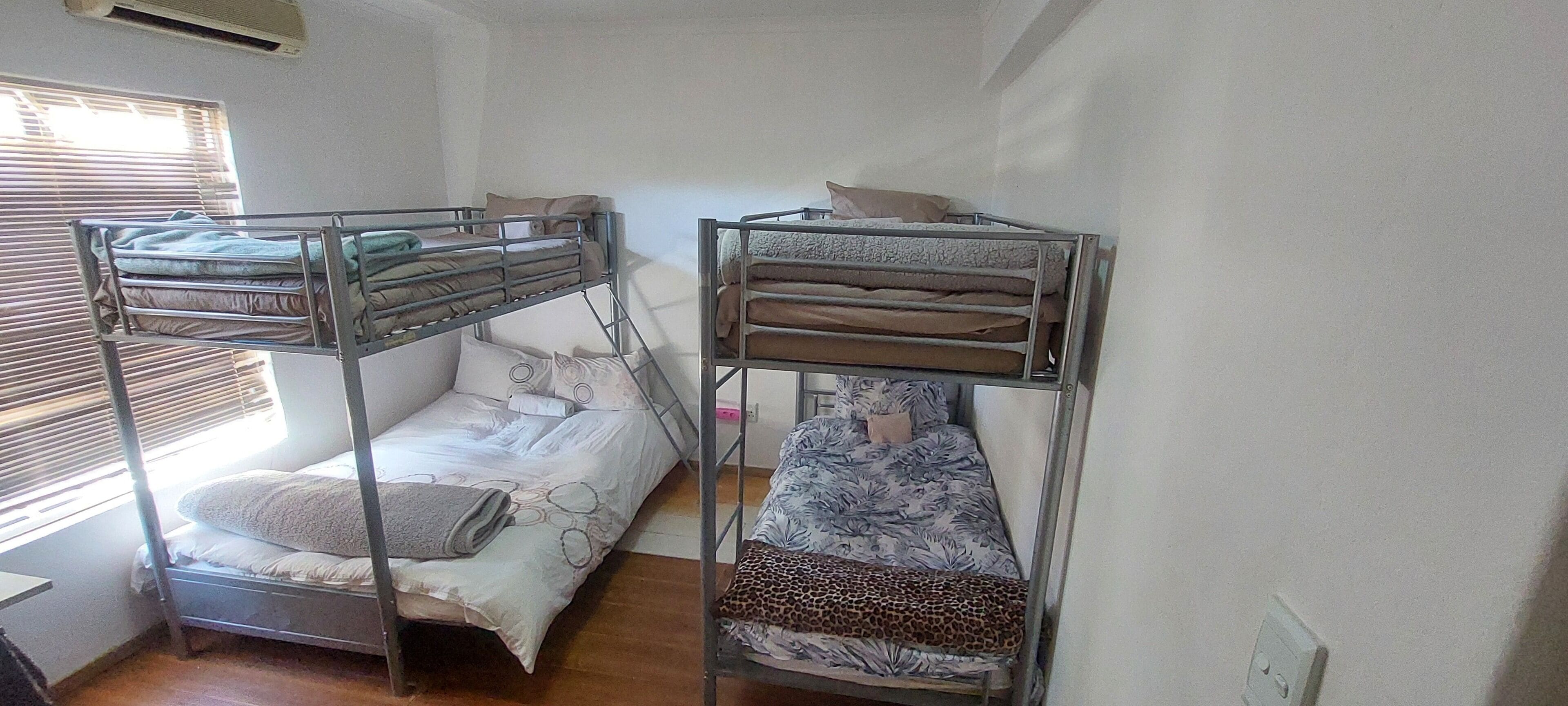 Een slaapzaal met twee stapelbedden, een raam met zonwering, en een witte muur.
